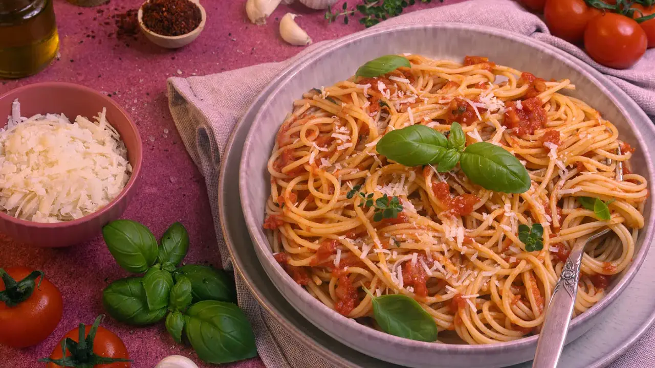 Ein Teller Spaghetti Napoli mit Tomatensauce, Basilikum und Parmesan. Daneben Zutaten wie Tomaten, Knoblauch und Olivenöl.
