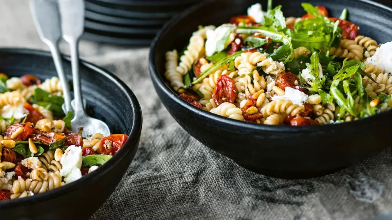 Zwei Schalen mit Nudelsalat mit getrockneten Tomaten, Pinienkernen und Rucola. Ein Löffel liegt in der vorderen Schale.