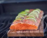 Ein saftiges Lachsfilet mit Limettenscheiben und Kräutern wird auf einem Holzbrett gegrillt.
