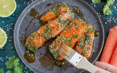 Vorbereitung für ein köstliches Gericht: Lachs marinieren mit Kräutern und Sesam. Eine Hand mit Pinsel verteilt die Marinade.