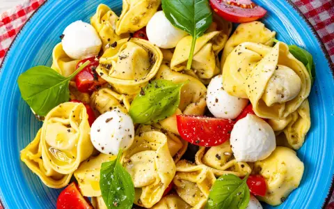 Ein bunter Tortellini Salat mit Mozzarella-Kugeln, Tomatenstücken und frischem Basilikum, serviert auf einem blauen Teller.