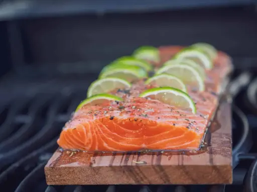 Ein saftiges Lachsfilet mit Limettenscheiben und Kräutern wird auf einem Holzbrett gegrillt.