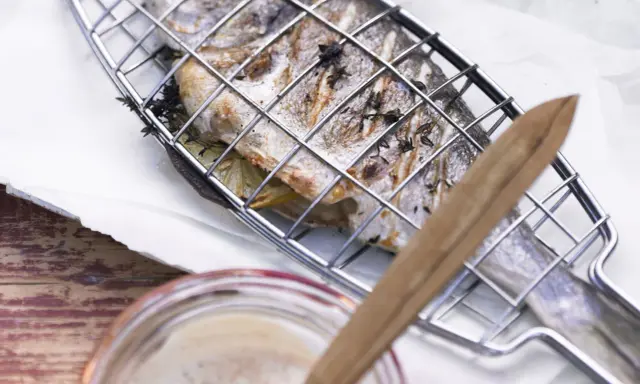 Ein ganzer Fisch liegt in einem Grillkorb, bereit zum Fisch grillen auf dem Elektrogrill. Kräuter und Zitronenscheiben sind im Fisch.