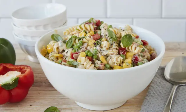 Ein bunter Nudelsalat ohne Mayonnaise mit Spiralnudeln, Erbsen, Mais, Paprika und frischem Basilikum in einer weißen Schüssel.