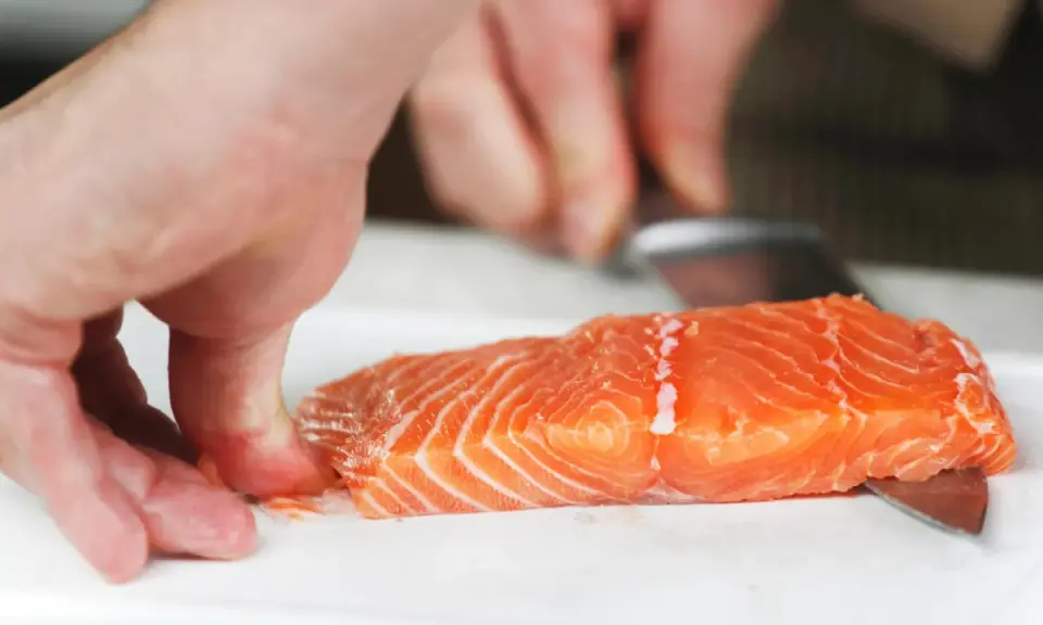 Hände halten ein Lachsfilet, während ein Messer die Haut vom Lachs entfernt.