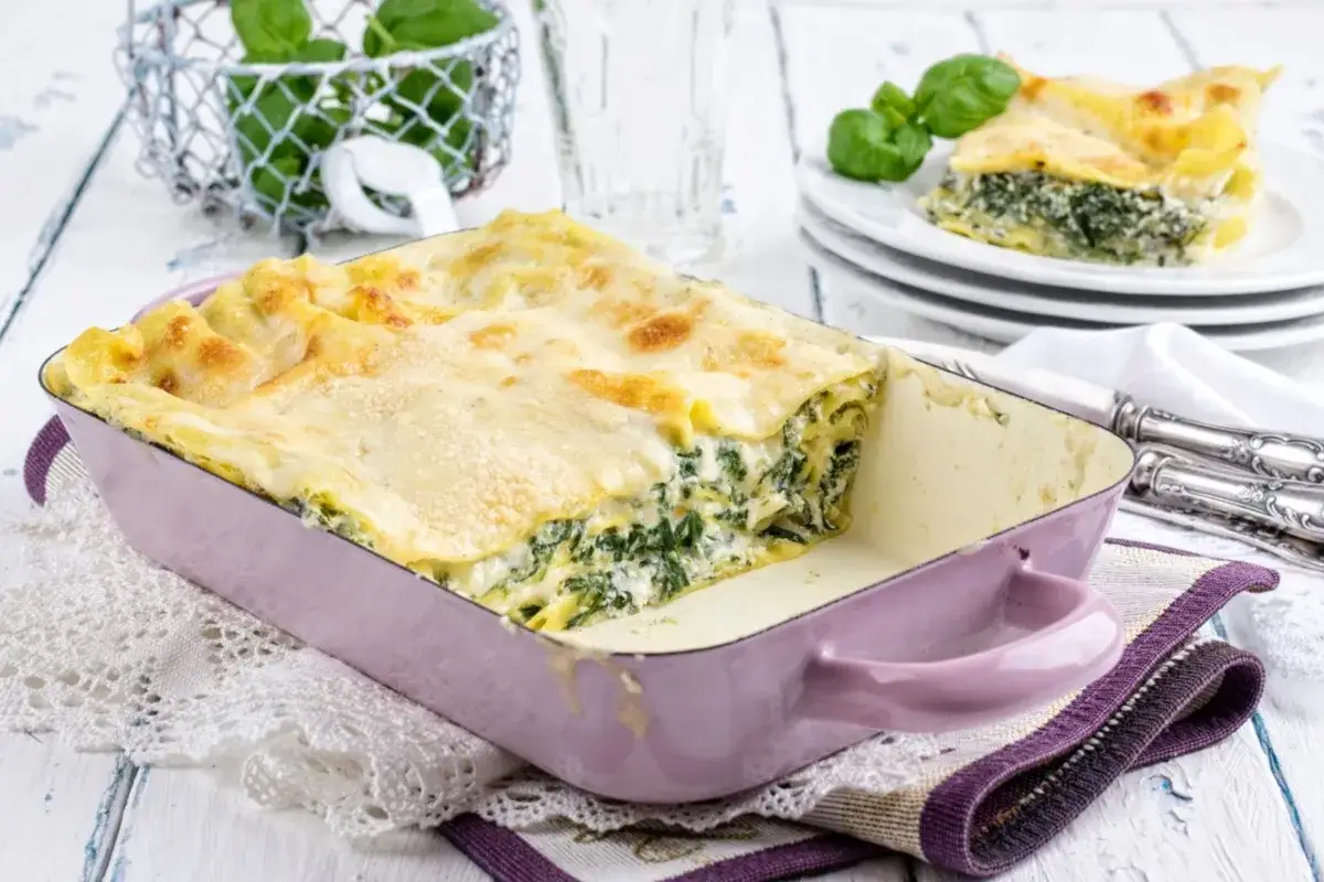 Herzhafte Spinat-Lasagne in einer lila Auflaufform, goldbraun &uuml;berbacken. Ein St&uuml;ck auf einem Teller daneben.