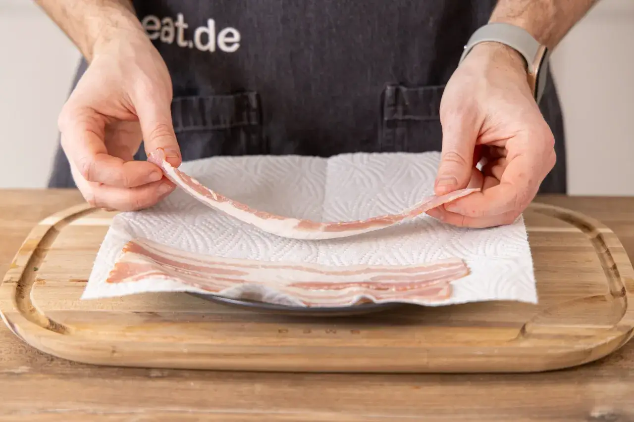 H&auml;nde legen rohen Bacon auf K&uuml;chenpapier, bereit f&uuml;r die Mikrowelle.