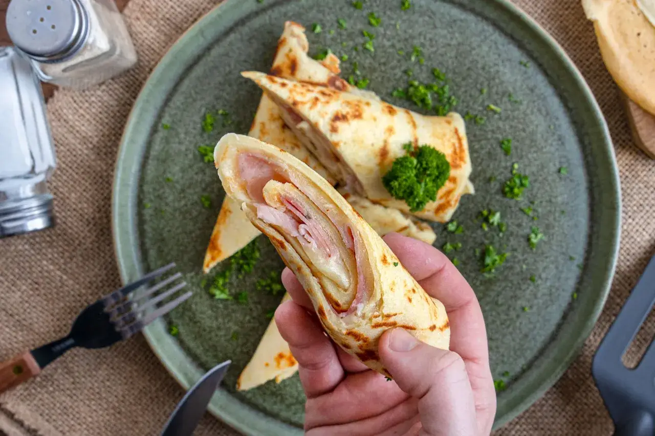 Eine Hand h&auml;lt einen herzhaften Pfannkuchen, gef&uuml;llt mit Schinken und K&auml;se. Auf dem Teller liegen weitere gerollte Pfannkuchen, garniert mit Petersilie.