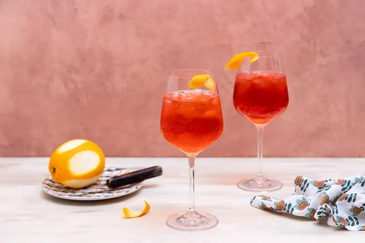Zwei erfrischende Campari Spritz mit Orangenzeste und Eis, daneben eine Orange und ein Stück Zimtstange.