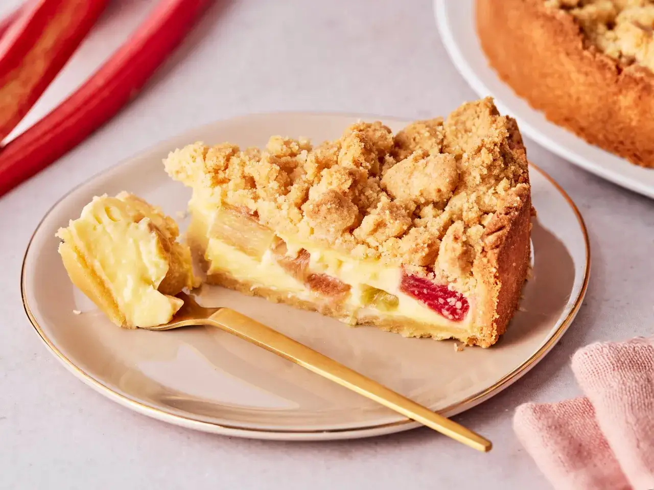 Ein saftiger Rhabarberkuchen mit Pudding und Streuseln auf einem Teller, daneben frische Rhabarberstangen.