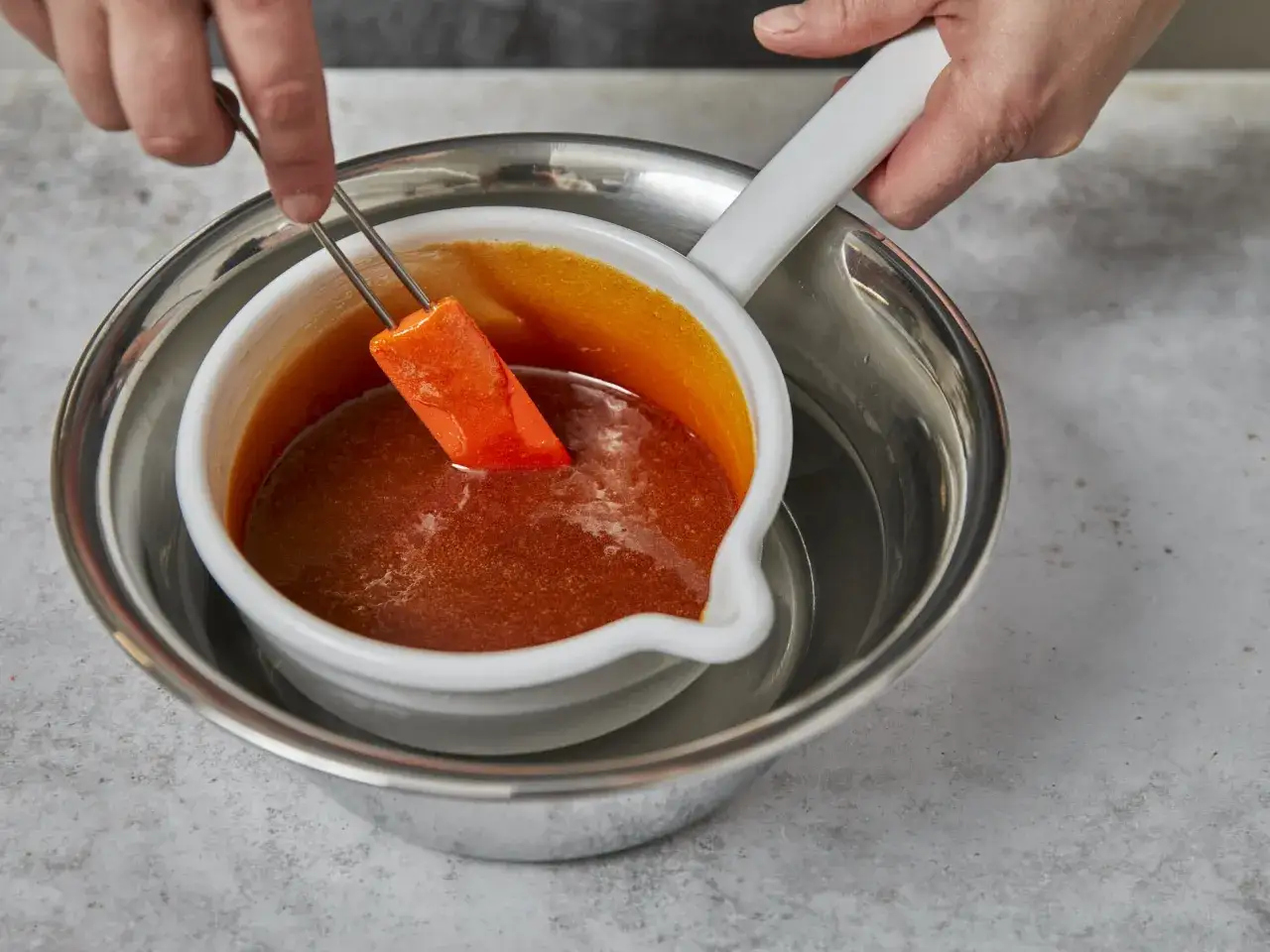 Ein St&uuml;ck orangefarbener Frucht wird mit einer Zange in eine Sch&uuml;ssel mit karamellisierendem Zucker getaucht.