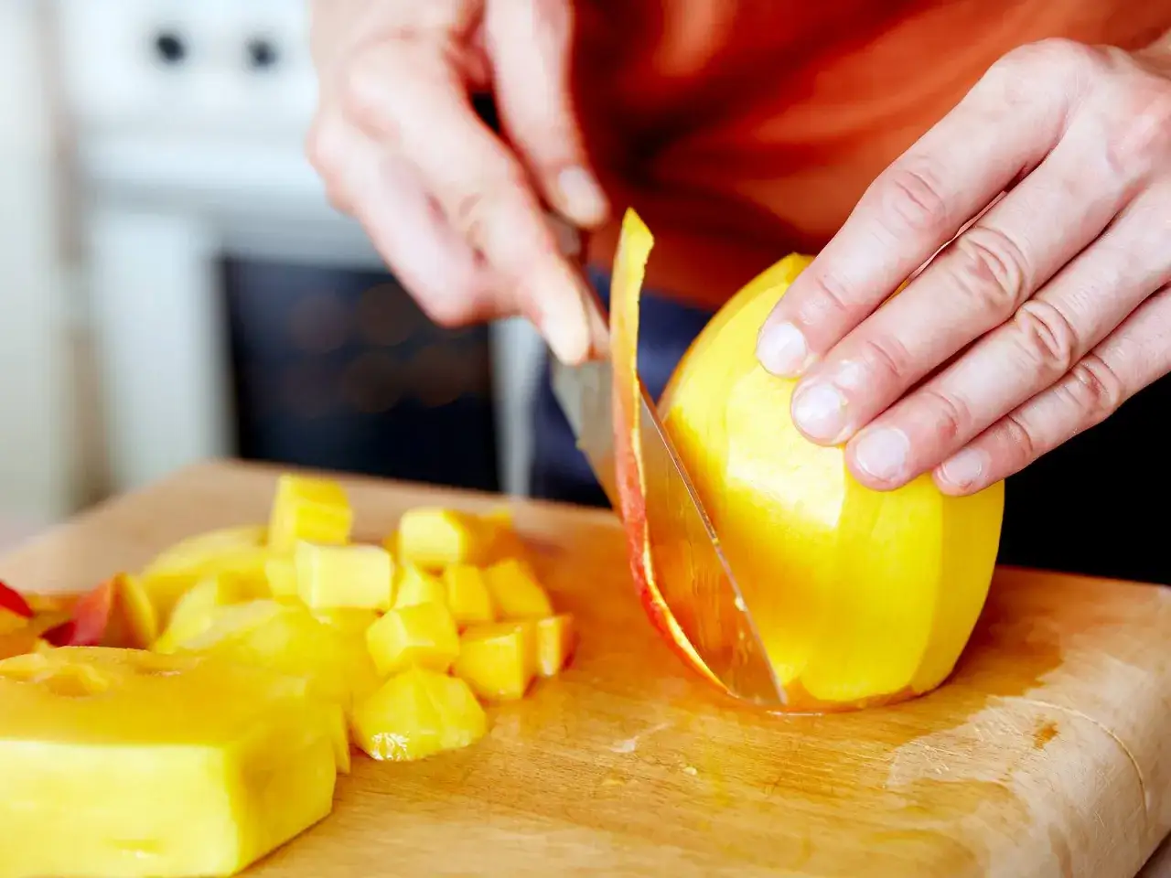 H&auml;nde schneiden eine reife Mango auf einem Holzbrett. Saftige, gelbe W&uuml;rfel liegen bereit.