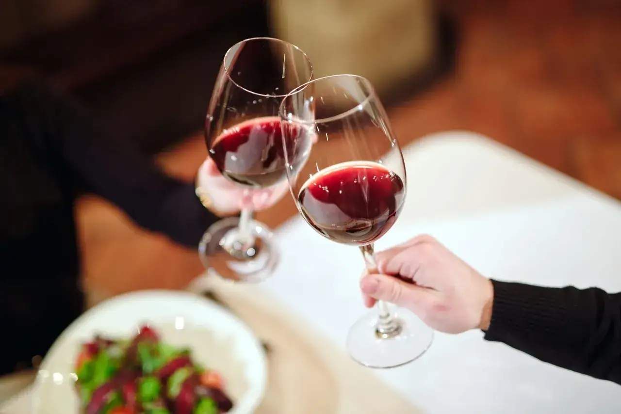 Zwei Gläser mit tiefrotem Wein rot stoßen an. Im Vordergrund ein Salat.