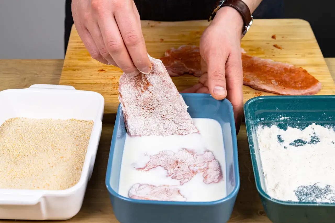 Fleisch wird in eine wei&szlig;e Fl&uuml;ssigkeit getaucht, um es f&uuml;r das Panieren ohne Ei vorzubereiten. Daneben liegen Paniermehl und Mehl.