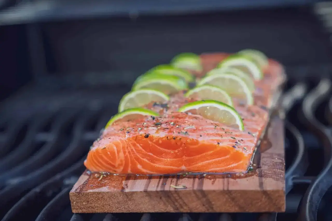 Ein saftiges Lachsfilet mit Limettenscheiben und Kr&auml;utern wird auf einem Zedernholzbrett gegrillt.