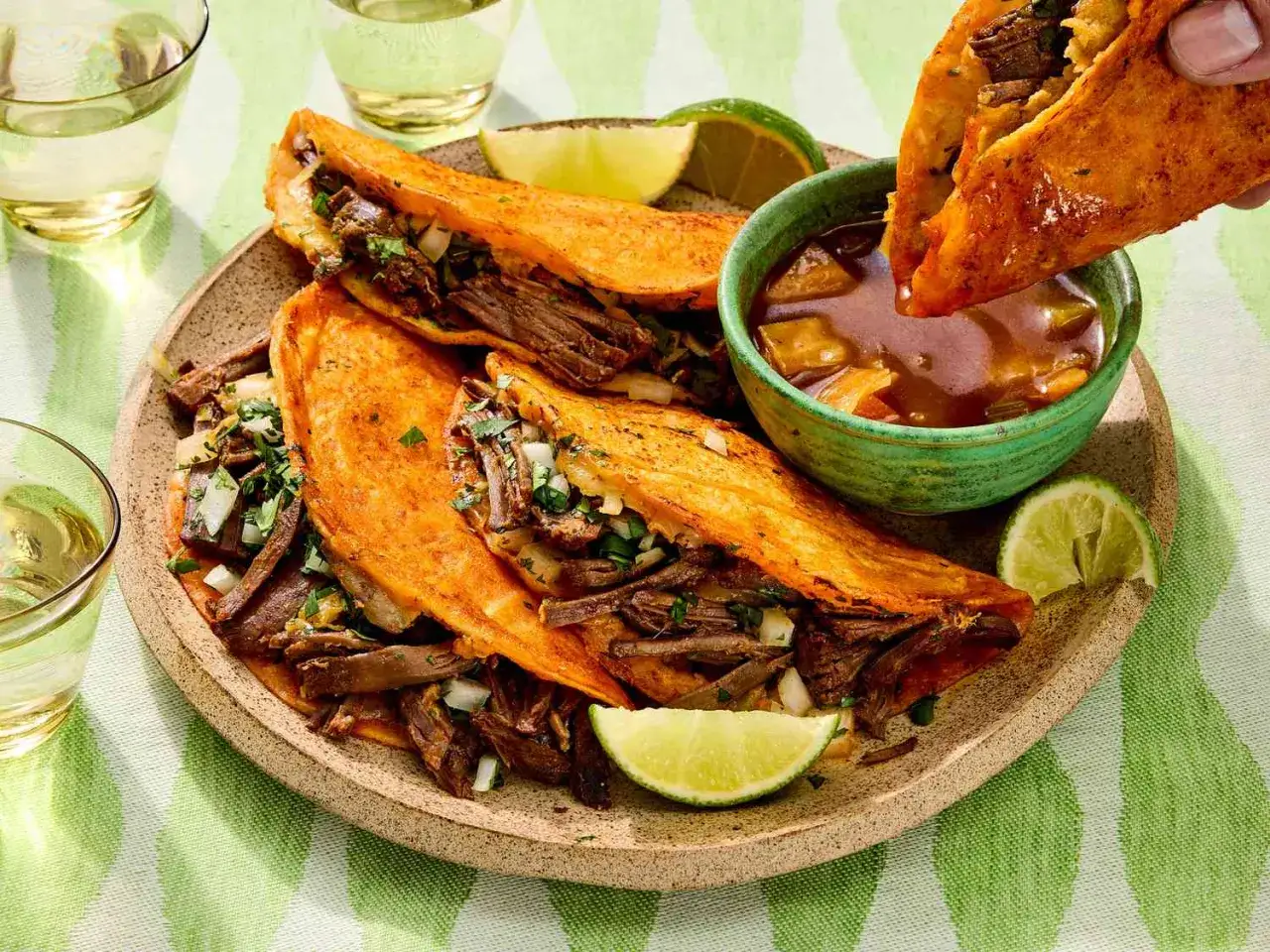Saftige birria tacos mit Rindfleisch, Zwiebeln und Koriander, zum Dippen in Consom&eacute;. Dazu Limettenspalten und Gl&auml;ser mit hellem Getr&auml;nk.