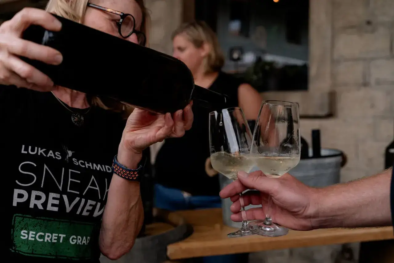 Eine Frau schenkt Scheurebe Wein in Gl&auml;ser ein. Ein Mann h&auml;lt zwei Gl&auml;ser mit dem hellgelben Wein.