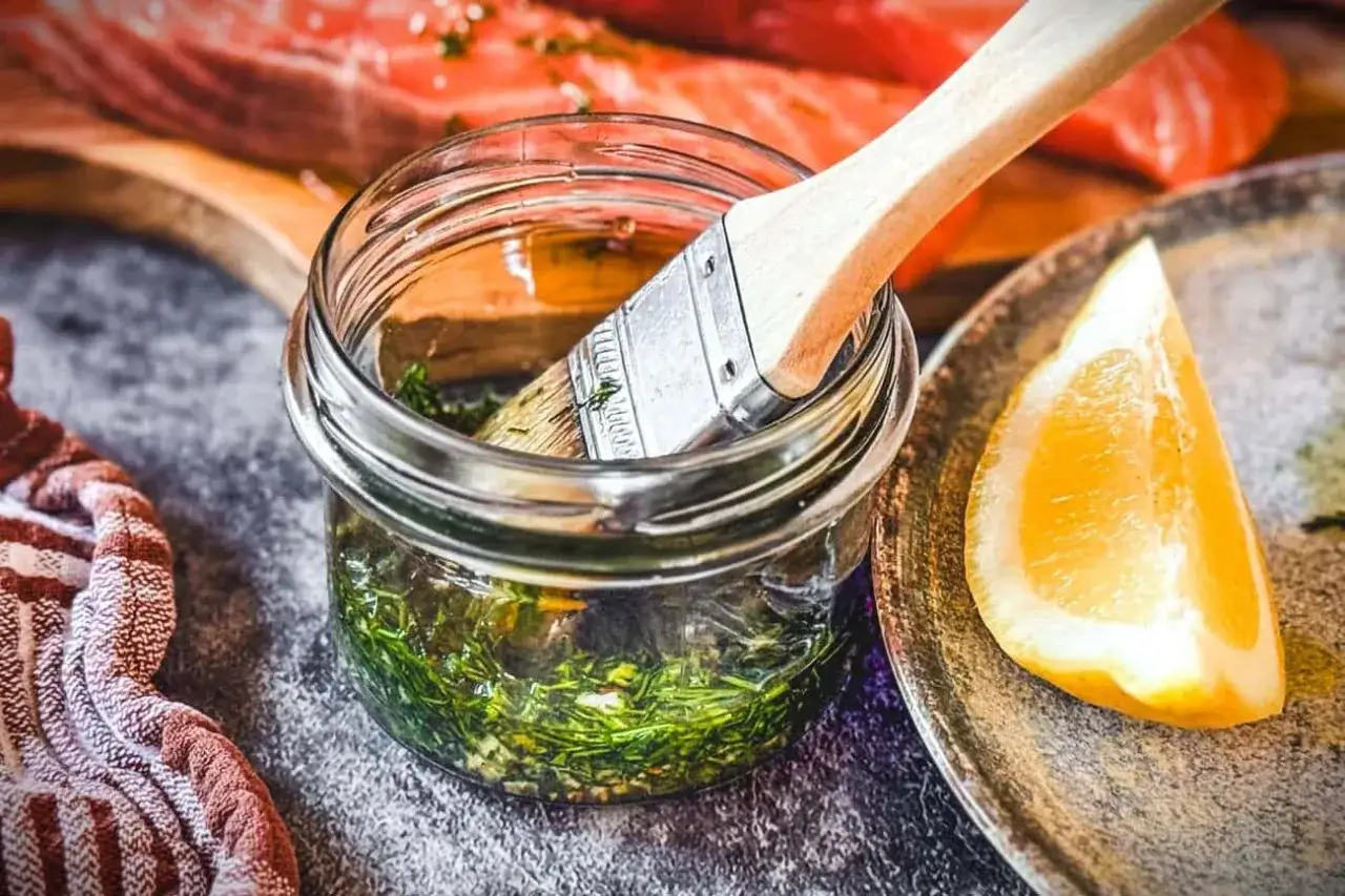 Marinade mit Kr&auml;utern und Zitrone bereit, um Lachs zu verfeinern. Ein Pinsel liegt in der Glasflasche.