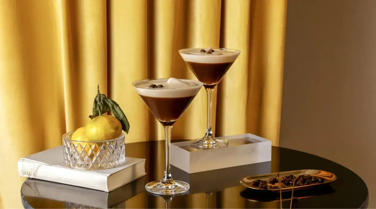 Zwei elegante Gl&auml;ser mit cremigem Espresso Martini alkoholfrei, garniert mit Kaffeebohnen. Daneben Zitrusfr&uuml;chte und Kaffeebohnen auf einem Tablett.