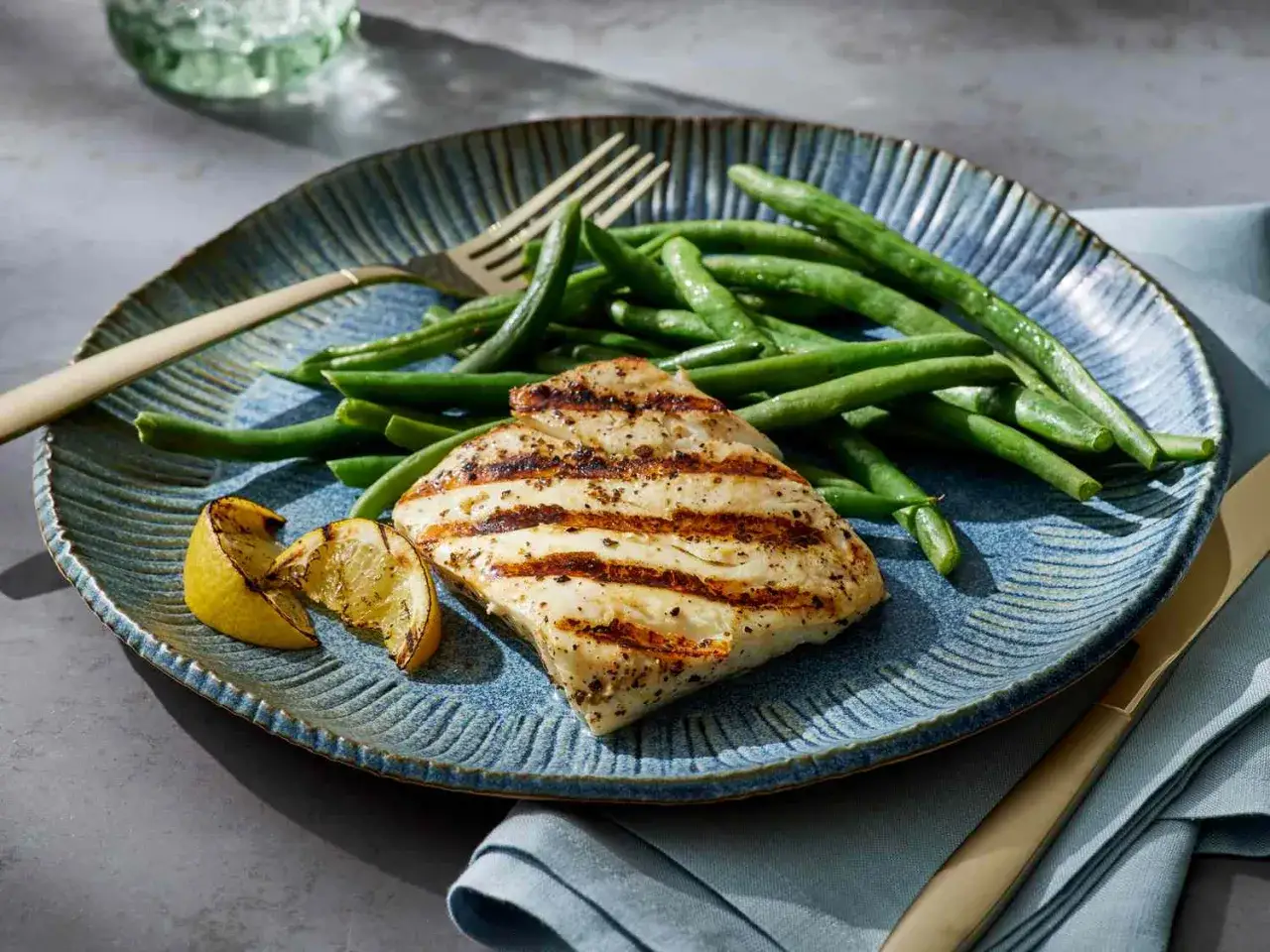 Perfekt gegrillter Fisch mit gr&uuml;nen Bohnen und Zitronenspalten auf einem blauen Teller. Ein k&ouml;stliches Gericht, das Lust auf Fisch grillen macht.