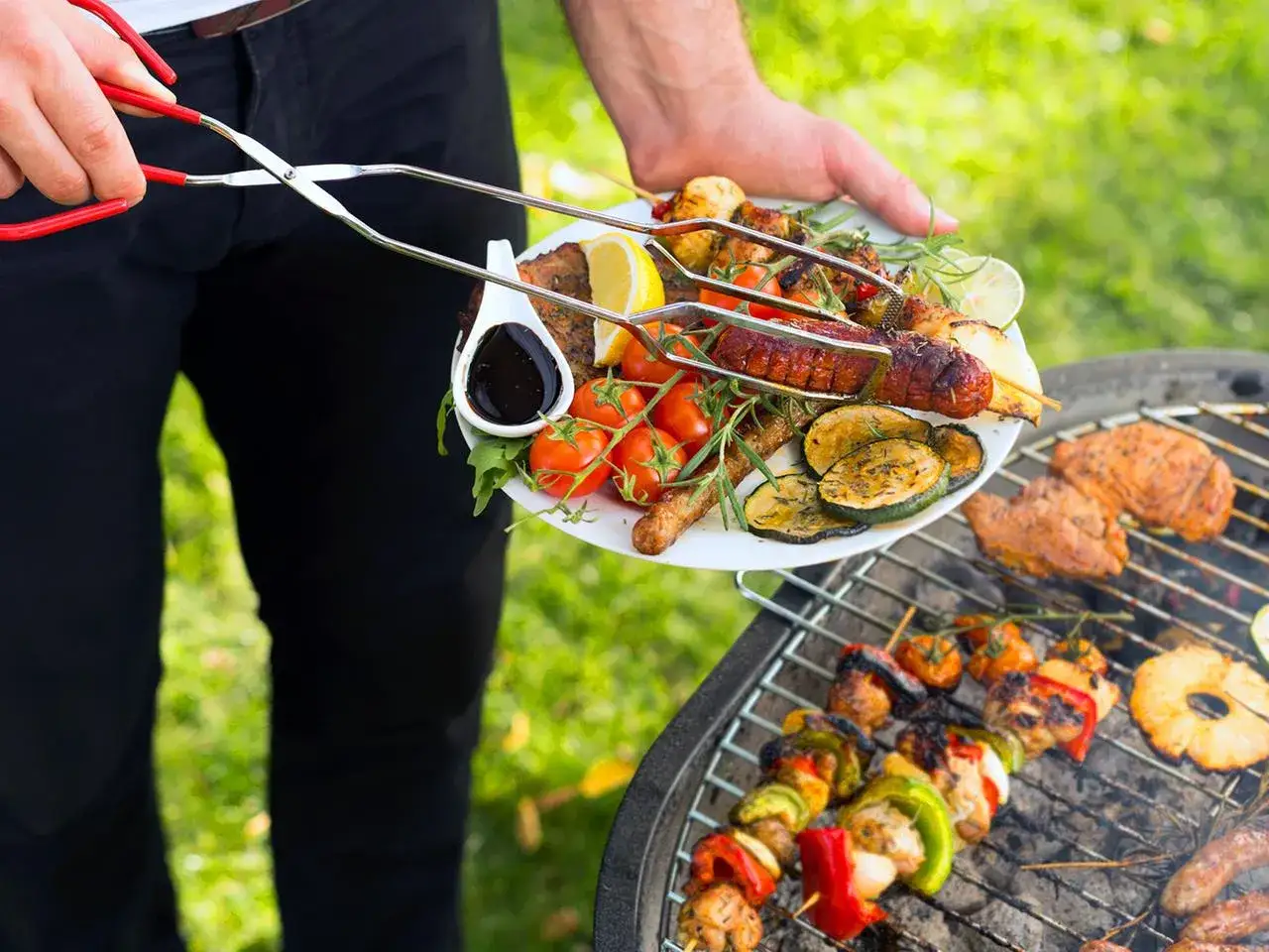 Leckere Grillideen: Ein Teller mit W&uuml;rstchen, H&auml;hnchenspie&szlig;en, Gem&uuml;se und Zitronenspalten wird vom Grill genommen.